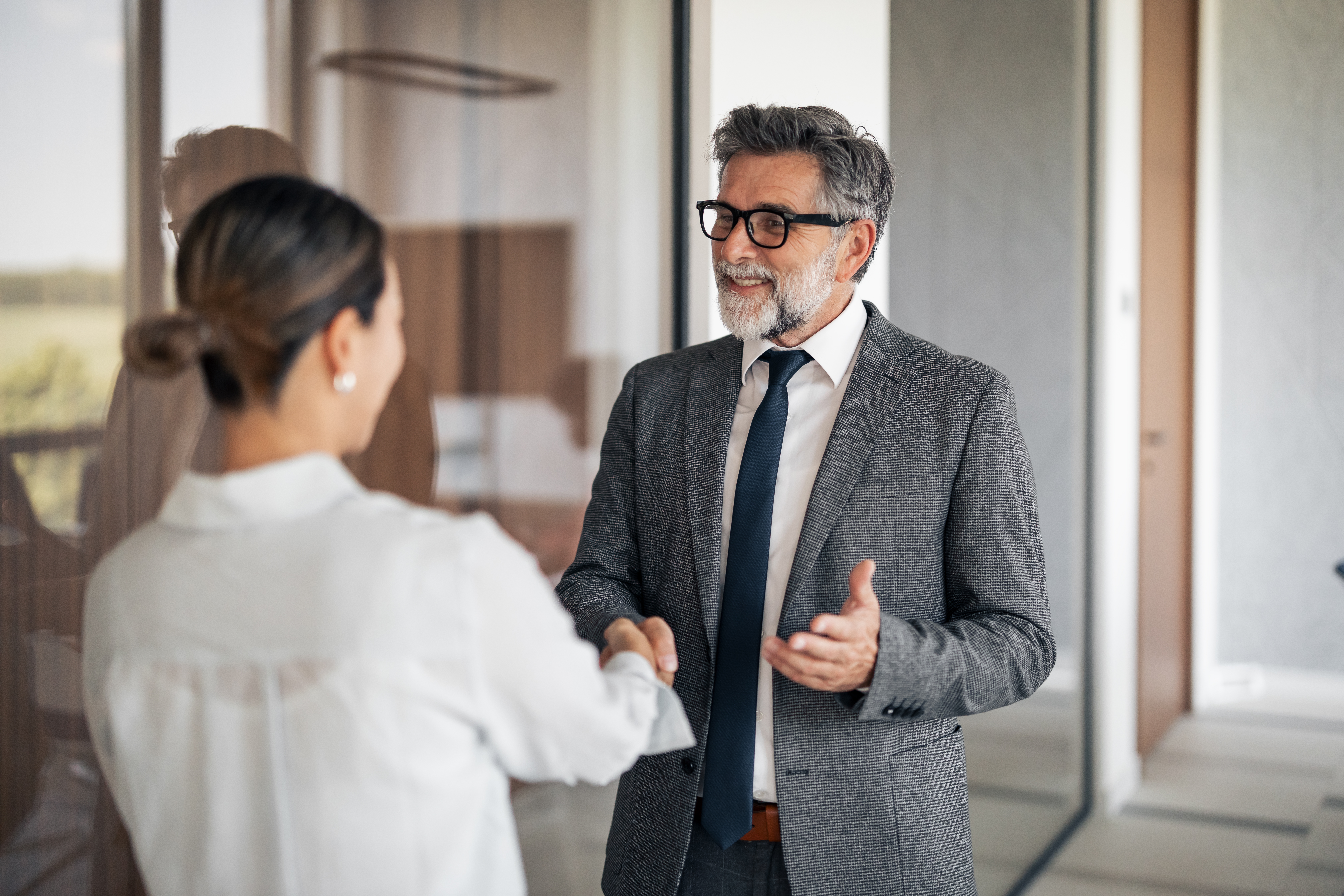 Two business professionals, a woman and a smiling mature adult man with a beard and eyeglasses, shaking hands in a modern office, confirming a partnership or successful deal